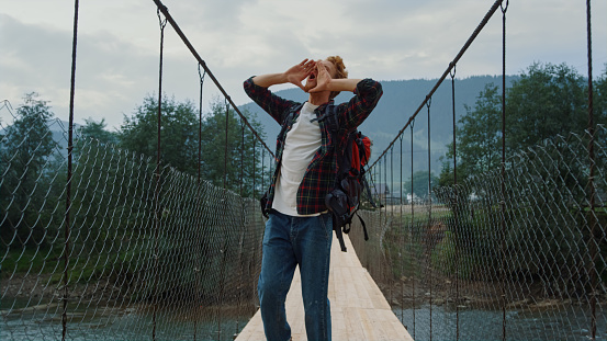Millennial backpacker run nature mountains. Cheerful man raise hands on travel close up. Emotional tourist scream feel freedom on river bridge. Carefree traveler celebrate enjoy hike. Active concept.