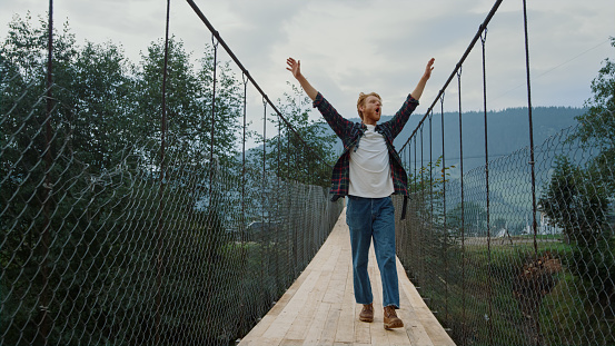 Carefree guy explore nature on mountains river bridge. Excited hiker walk outdoors on active vacation. Happy traveler enjoy freedom on tourism activity. Redhead backpacker trekking. Happiness concept.