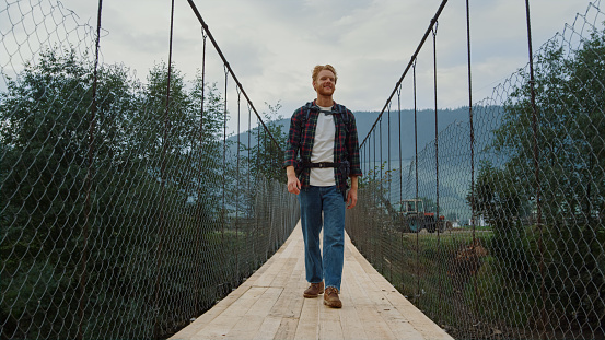 Traveling man walk bridge in countryside mountains. Smiling guy explore nature. Positive tourist wear backpack on active hike sport. Young hipster spend weekend outdoors. Travel leisure trip concept.