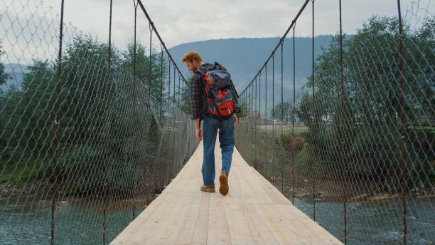 Hiker walk river bridge at mountains landscape nature. Back view backpacker enjoy tourism in green forest. Unknown traveler trekking outside. Young redhead tourist travel on vacation. Active concept.