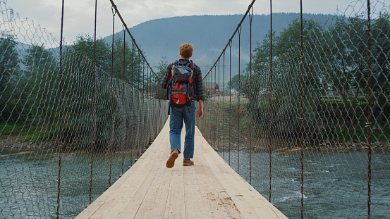 Curious backpacker traveling nature in mountains. Tourist walk on river bridge back view. Redhead guy enjoy active tourism at summer holiday vacation. Unknown hiker look around nature. Travel concept.