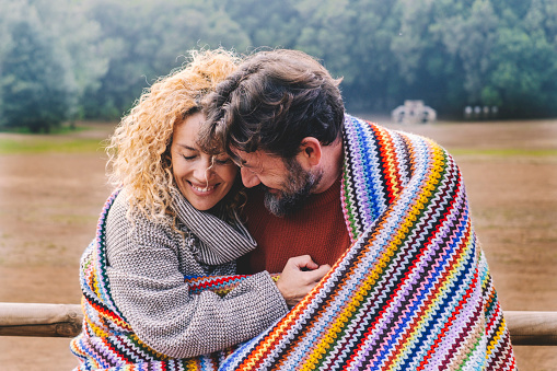 Romantic adult couple in love hug with tenderness under a colorful wool cover in outdoor park. Concept of winter leisure activity for man and woman in relationship. People embracing and enjoying