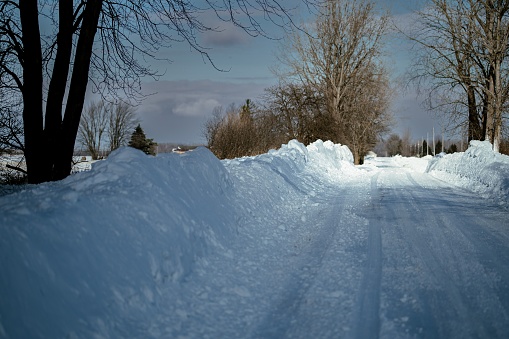 A rural road with plowed snow piles on the roadsides, and leafless trees on the way in Michigan, USA
