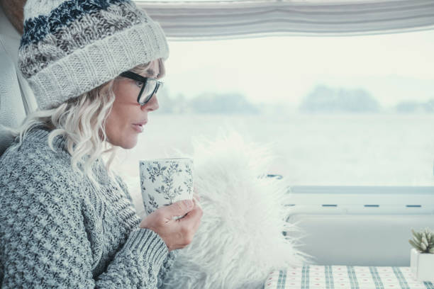 istock Side view of attractive lady with white hair and clothes drinking from a cup inside her camper van during winter season. Bright image of serene woman. Portrait of female people in inside leisure 1453577041