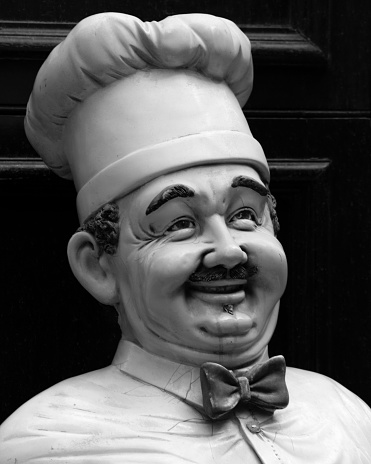 A closeup shot of a male cook statue dressed in uniform with a mustache and a big smile in greyscale