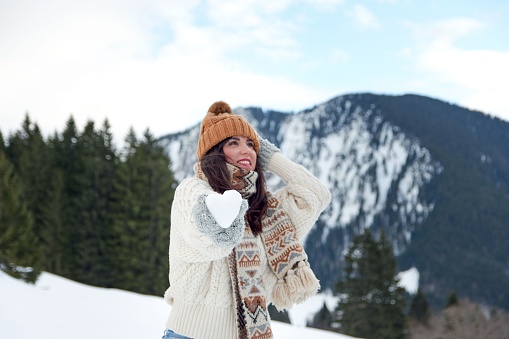 A female tourist with a scarf and a winter hat holding a heart-shaped snowball towards the camera