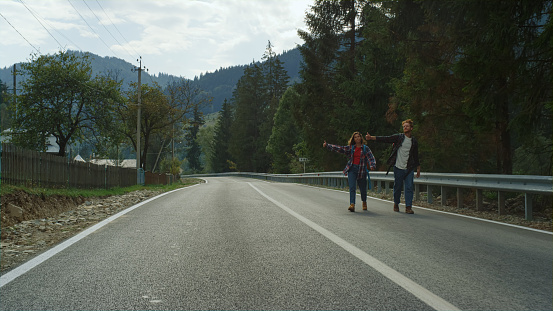 Happy tourists hitchhiking car on highway. Smiling friends wave hands in mountains. Couple have fun on forest hike. Positive lovers spend time together on weekend travel activity. Leisure concept.