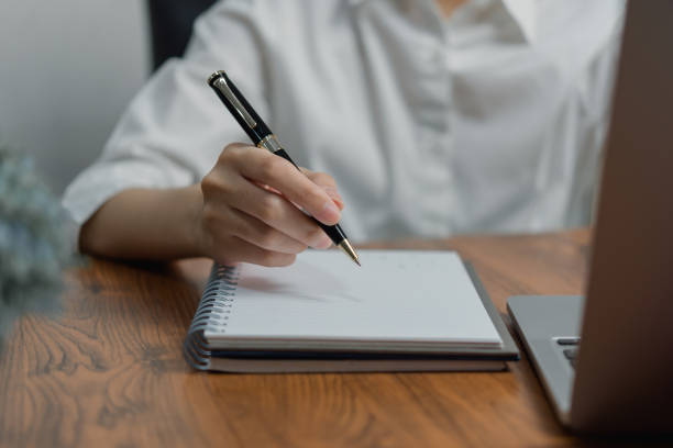 istock woman hand holding a pen and writing information in a notebook working with laptop computer.Education learning, journal diary. 1453608754