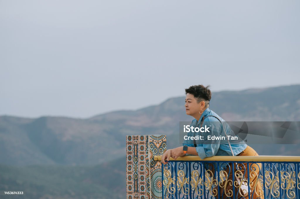 Asian Chinese female tourist leaning on fence looking away admiring Forêt Toufliht, Morocco rural mountain range scenery 45-49 Years Stock Photo
