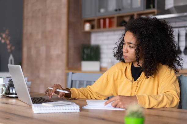 istock An african-american young woman is using laptop computer for remote work or studying online, she takes notes watching webinars or classes at kitchen table at home 1453620997