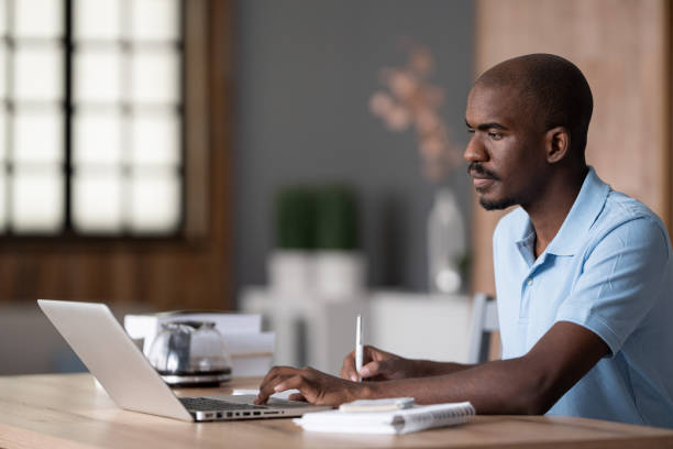 istock Online Education. Serious african man studying and working online, watching webinar on a laptop and taking notes. African man on a distance learning, sitting at kitchen table at home 1453620998