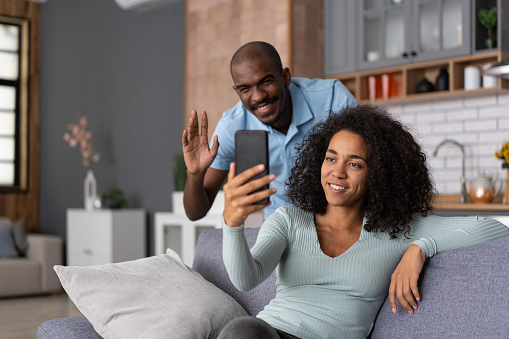 Remote Communication. African American man and woman waving palms at a mobile phone, sitting on the sofa in kitchen room interior. Cheerful black couple having video call via cellphone, staying at home
