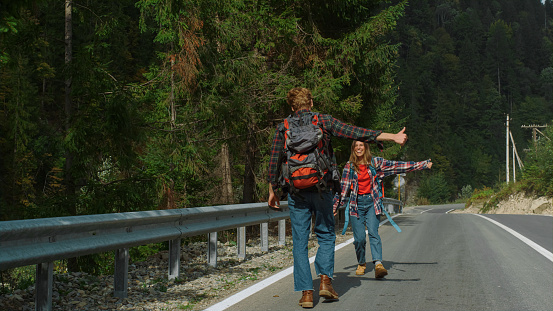 Overjoyed hitchhikers waving hands on mountain road. Couple jump on highway outdoors. Smiling tourists hiking car on nature roadside. Two happy friends showing thumbs up in forest. Travel fun concept.