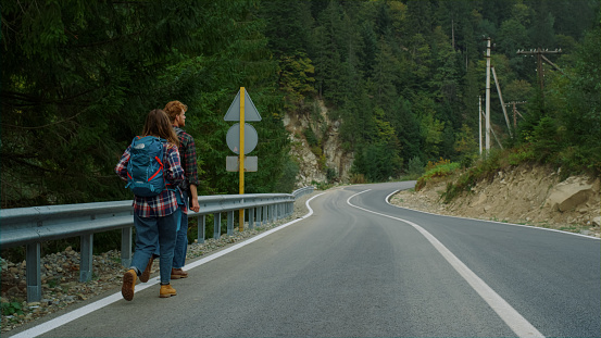 Couple hitchhikers walk mountains. Two travelers trekking on roadside highway back view. Friends exploring nature on vacation holiday. Young tourists wear backpacks for travel. Holiday tourism concept