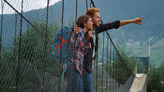 Two friends exploring mountains on summer holiday. Travelers rest on hitchhike trekking adventure. Smiling couple talking on river bridge. Hikers pointing finger on landscape view. Leisure concept.