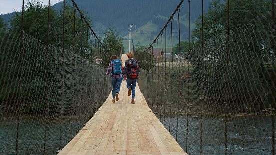 Couple run mountains bridge on nature river. Excited travelers have fun outside back view. Unknown lovers hitchhiking on weekend vacation. Hikers enjoying tourism in summer holiday. Leisure concept.