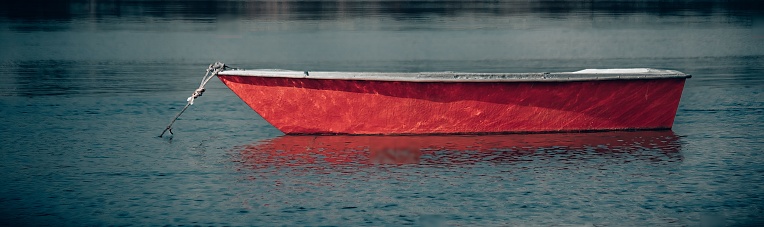 A small catboat with a red sail covering rests, moored in Quissett Harbor, in Falmouth, Massachusetts.