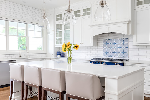 A luxury white kitchen with bar stools sitting at a large island, glass lights hanging from the ceiling, and a beautiful tiled backsplash.