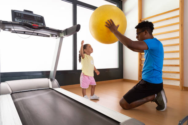 Side view of African American dad and mixed race child holding yellow fit ball in raised arms during fitness workout in gym