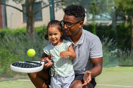 African American dad teaching mixed race child to bounce ball on racket during paddle tennis lesson on court on summer day