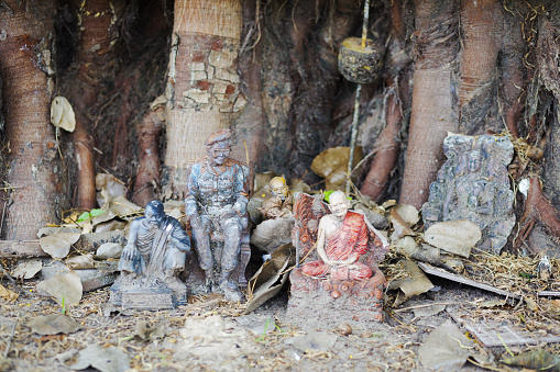 Thai buddhist shrine with several figurines including princess Srinagarindra placed at a tropical tree in rural area of Nong Khai province. Princess statue is between thai monks and buddha statues. Princess Srinagarindra is also called princess of humanity