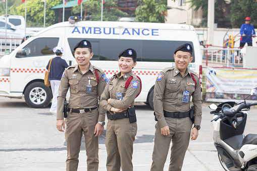 Capture of three cheerful thai tourist police officers in front of ambulance van on  promenade in Nong Khai in early morning and sunrise at Mekong river in North Thailand. There are two men and one woman. In background are a few people and preparations for a local cycling event