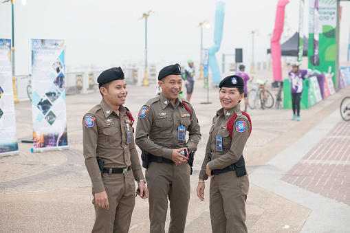 Three cheerful thai tourist police officers are standing  on  promenade in Nong Khai in early morning and sunrise at Mekong river in North Thailand. There are two men and one woman. In background are a few people and preparations for a local cycling event