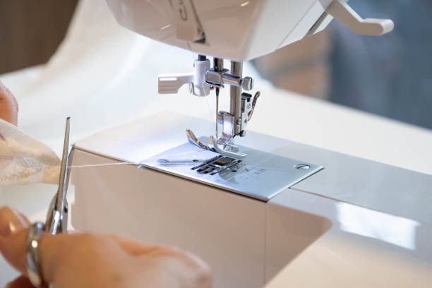 istock Cropped female hands sewing fabric in manufacturing machine at workplace cutting thread with scissors. 1453644264