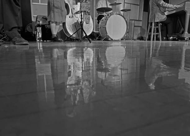 View below the heads of band members playing on a stage.   In black and white.\nPiano, drums, and bass players all unrecognizable since no faces or heads are pictured.  Reflection of band on polished, stage floor.
