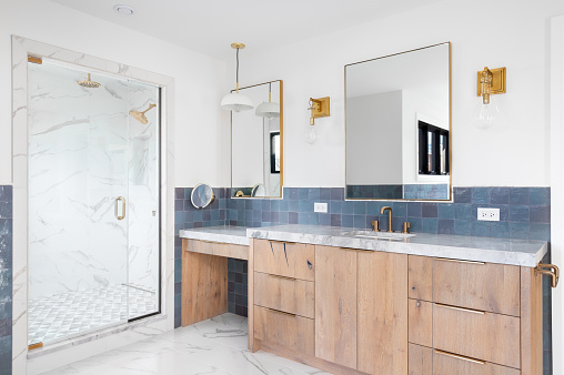 A luxurious bathroom with a wood vanity cabinet, blue tiles on the walls, gold accented lights and mirrors, and a large walk-in shower lined with marble tiles.