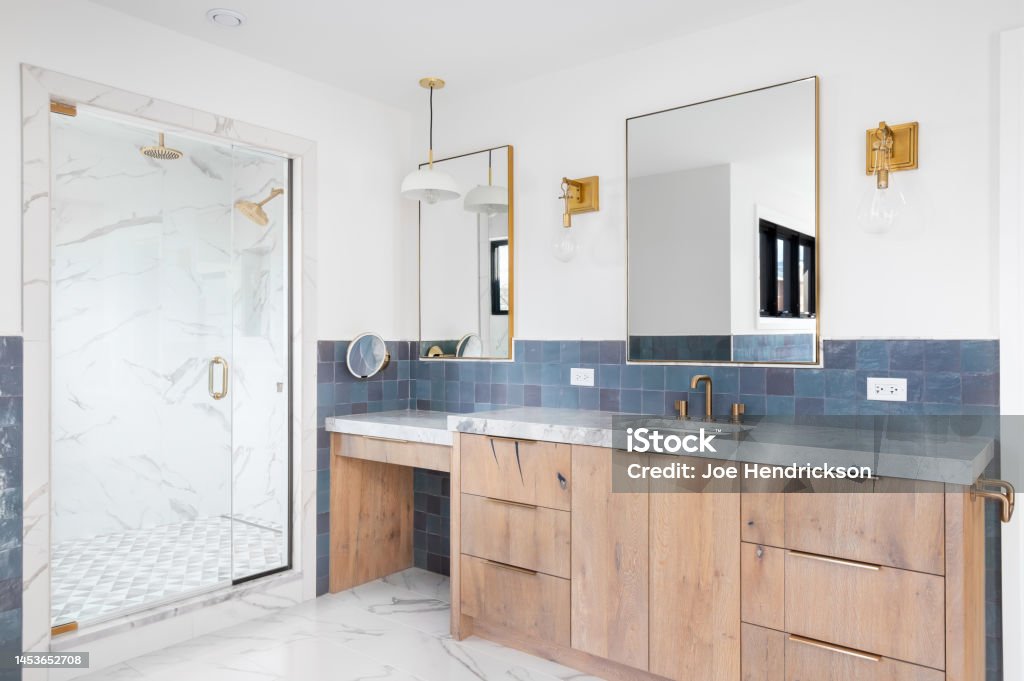 A bathroom with a wood cabinet, gold accents, and a marble shower. A luxurious bathroom with a wood vanity cabinet, blue tiles on the walls, gold accented lights and mirrors, and a large walk-in shower lined with marble tiles. Bathroom Stock Photo