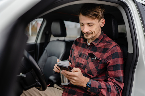 One man caucasian male with mustaches sit in the car take a brake on road-trip hold cup of coffee use mobile phone smartphone for sms texting or navigation app real people copy space