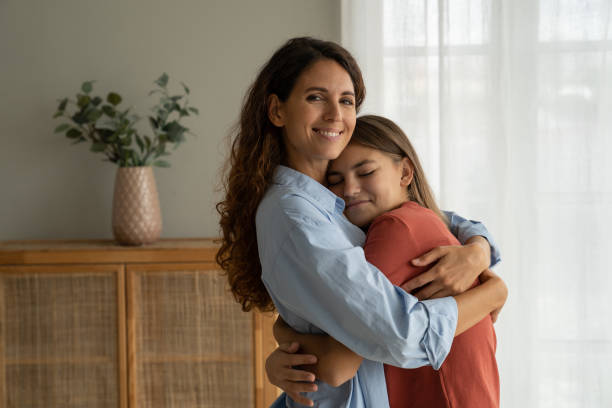 Happy family young mother and teen girl daughter cuddling hugging at home Young loving mother hugging cuddling with teen girl daughter at home and smiling at camera, expressing unconditional love. Mother-daughter friendship, happy motherhood, parenting of adolescent candid bonding connection togetherness stock pictures, royalty-free photos & images