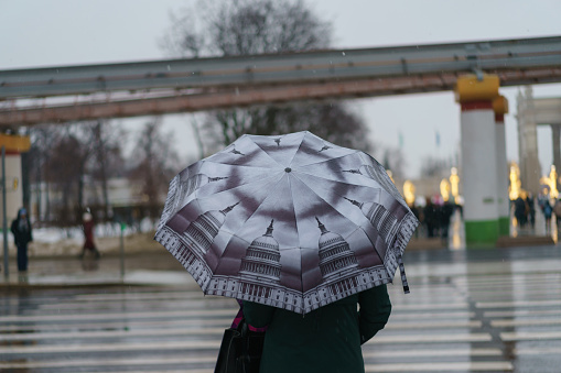 Photography of the people on the city street during rain with snow. Rainy gloomy day Woman stands in front of the motorway, to cross the street. Back view