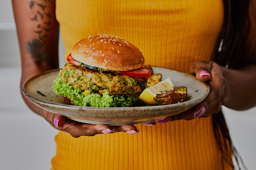 A multiracial woman in a casual yellow dress, holding a healthy vegan hamburger on a handmade ceramic plate, made of zucchini, green pea, seasoning, herbs and spices, close up