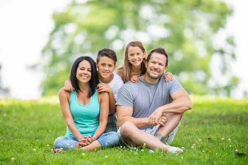 A young family of four sit outside in the grass on a sunny summers day as they pose for a portrait.  They are each dressed casually and are smiling as they huddle in closely to one another.
