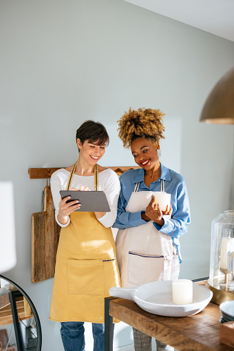 Smiling African-American woman holding candles while cheerful caucasian woman holding and showing digital tablet to her.