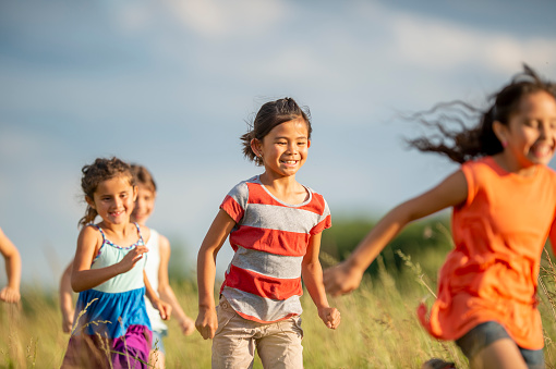 A large group of school aged children are seen running through a field of tall grass on a sunny summers day.  They are each dressed casually and are smiling as they enjoy the fresh air and each others company.