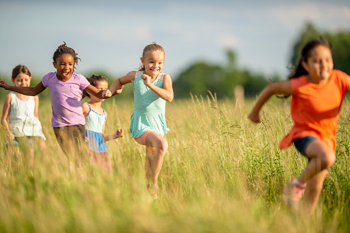 A large group of school aged children are seen running through a field of tall grass on a sunny summers day.  They are each dressed casually and are smiling as they enjoy the fresh air and each others company.