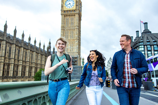 Goofy adult caucasian friends having fun while exploring London's landmarks. They are walking by the Palace of Westminster. They are all looking at each other while laughing and rushing through the city. In the background there is the Elizabeth Tower. They are carrying backpacks and a camera.