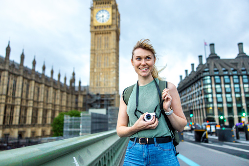 A portrait of an attractive caucasian young adult female tourist looking away from the camera during their London trip. She is standing on a bridge near the Palace of Westminster. In the background there is the famous clock tower Big Ben. The tourist is smiling and looks happy. Her blonde hair is tied in a ponytail. She has a backpack on her back.