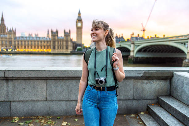 Caucasian Young Adult Female Tourist Smiling As She Is Exploring London's Landmarks Attractive young adult caucasian female tourist smiling while walking away from the fence by the river Thames in London. She is exploring the city alone. There is a camera hanging around her neck. Behind her there is the beautiful Elizabeth Tower and The Palace of Westminster. The sky is beautiful since the sun is setting. westminster bridge stock pictures, royalty-free photos & images