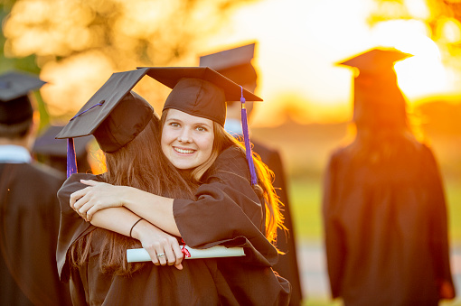 Two young female graduates take a moment to embrace and congratulate one other after graduation.  They are both wearing caps and gowns and are holding their diplomas in hand.