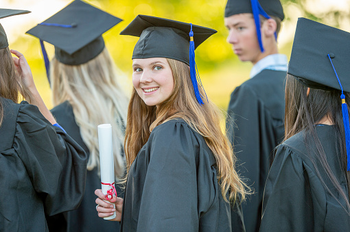 Back view of brunette lady in graduation costume raising her diploma up, unrecognizable female student posing at university campus, celebrating graduation, showing her success, copy space