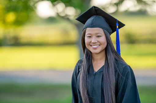 Beautiful mixed race Japanese Mexican young woman college graduate portrait wearing cap and gown with diploma isolated on white background