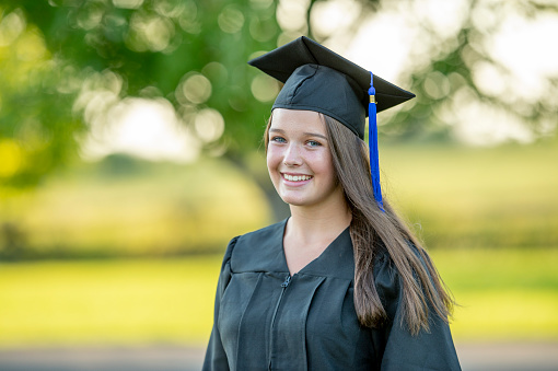 A young female graduate stands out side as she poses for a portrait.  She is wearing a gown and cap and smiling for the camera.