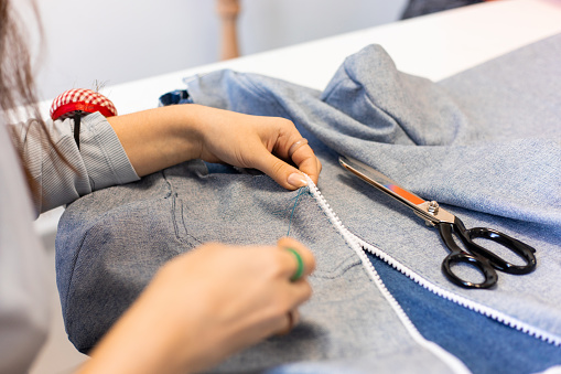 Close-up, female hands sewing on fabric with zipper using needle and thread. Scissors are on the right. The woman has pins in her arm. Fashion designer.