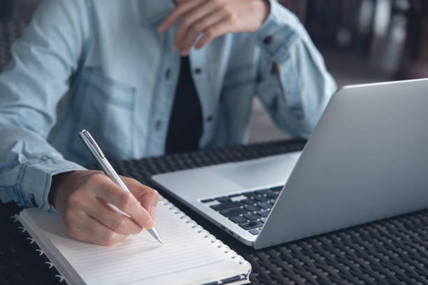 istock Online class, Student writing on notebook while study at home. Young adult woman doing online lesson by using laptop computer 1453685483