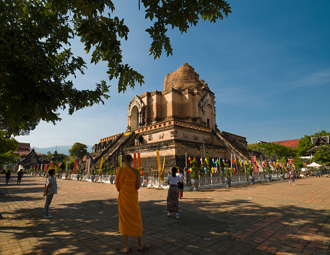 Rear view of sitting  of adult and mature thai women at buddhist ceremony outside of a temple captured in Chiang Rai province in area of Chiang Khong. Women are gathering together for a local buddhist ceremony with a male musician playing classical string instrument and prayers. Women are sitting  on a plaid on floor around a buddhist flower decor. In background is the local temple. One woman is walking in scene