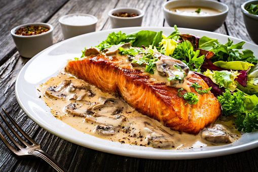 Fried salmon steak with mushrooms in sauce and leafy greens on wooden table